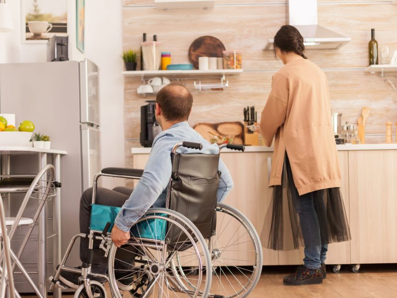Man with walking handicap in wheelchair looking at wife how she's cooking. Disabled paralyzed handicapped man with walking disability integrating after an accident.